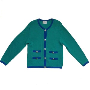May include: Teal green cardigan with a blue trim and silver buttons. The long-sleeved sweater has two front pockets with silver button details. The collar and cuffs are trimmed with blue. The cardigan is buttoned up.