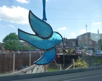 May include: A stained glass blue bird ornament with a dark border, hanging from a blue ribbon. The bird is holding a small charm. The background shows a residential area with houses and trees.