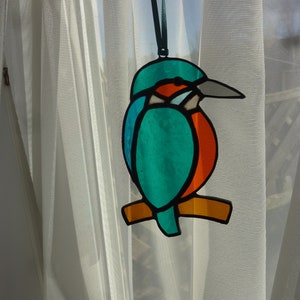 May include: A stained glass bird ornament hanging from a white sheer curtain. The bird is blue, green, orange, and red.
