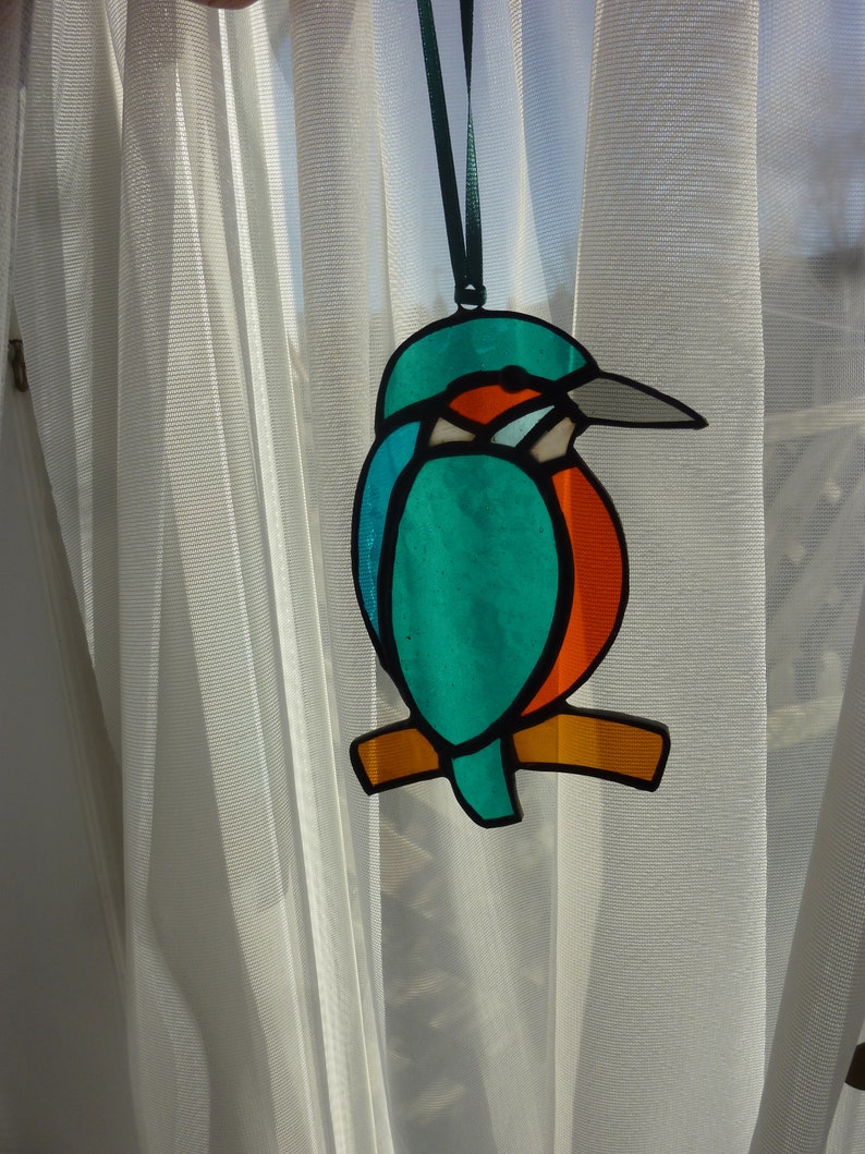 May include: A stained glass bird ornament hanging from a white sheer curtain. The bird is blue, green, orange, and red.