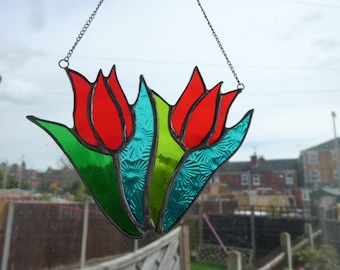 May include: A stained glass window hanging featuring a design of three red tulips with green leaves and blue accents.