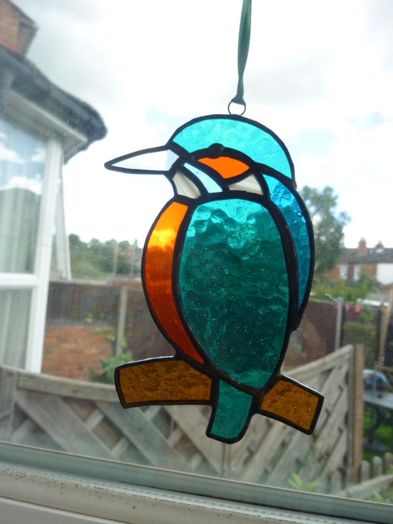 May include: A stained glass window hanging ornament depicting a blue and orange kingfisher bird with a brown branch.