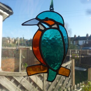 May include: A stained glass hanging ornament of a kingfisher bird. The bird is blue, green, orange, and white. The bird is hanging from a green ribbon.