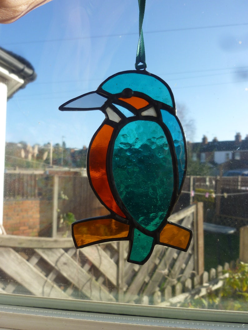 May include: A stained glass hanging ornament of a kingfisher bird. The bird is blue, green, orange, and white. The bird is hanging from a green ribbon.