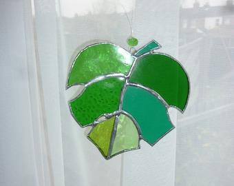 May include: A stained glass leaf suncatcher with a variety of green hues, including textured and smooth glass panels. The leaf is outlined with a silver-colored metal and has a small green bead at the top for hanging. The suncatcher is displayed in front of a sheer white curtain.