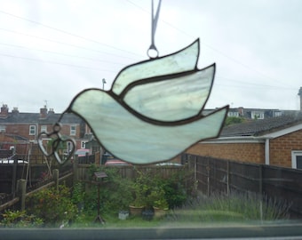 May include: A stained glass suncatcher in the shape of a flying dove with iridescent white and pale blue glass panels held together by dark metal solder lines, featuring two small dangling metal heart charms hanging from the bird's beak.