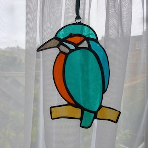 May include: A stained glass hanging ornament depicting a blue and orange kingfisher perched on a branch. The bird has a long, thin beak and a bright blue body with orange accents.