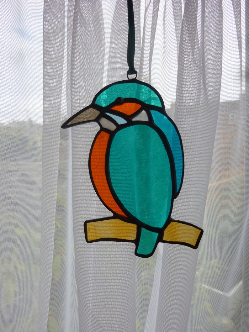 May include: A stained glass hanging ornament depicting a blue and orange kingfisher perched on a branch. The bird has a long, thin beak and a bright blue body with orange accents.