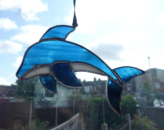 Puede incluir: Un delfín de vidriera en tonos azules y blancos, con borde negro. El delfín está suspendido por un cordón negro sobre un fondo de cielo nublado y zona residencial. Una pieza decorativa.