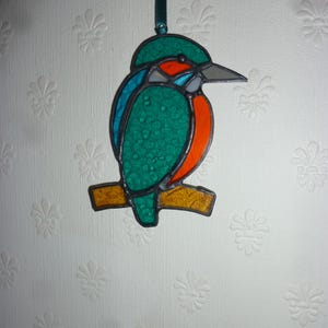 May include: A stained glass ornament of a kingfisher perched on a branch. The bird is green, blue, and orange with a long beak.