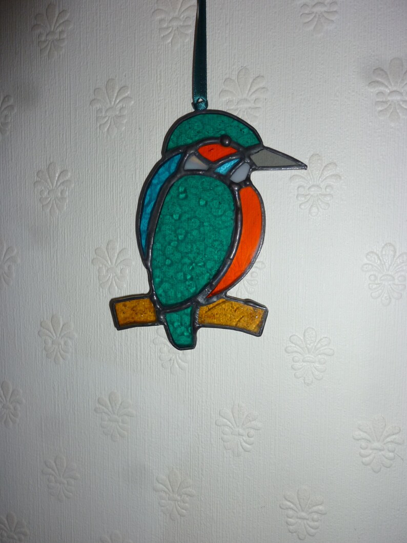 May include: A stained glass ornament of a kingfisher perched on a branch. The bird is green, blue, and orange with a long beak.