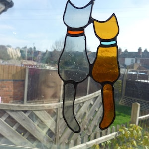 Puede incluir: Dos gatos de vidrio coloreado colgados en una ventana. El gato de la izquierda es gris con un collar naranja y el gato de la derecha es amarillo con un collar azul.