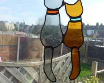 May include: Two stained glass cats hanging in a window. The cat on the left is gray with an orange collar and the cat on the right is yellow with a blue collar.