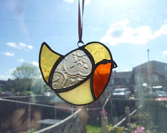 May include: Stained glass bird suncatcher with yellow, orange, and clear textured glass. The bird is hanging from a brown ribbon. The suncatcher is a decorative item for windows.