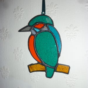 May include: A stained glass ornament of a kingfisher perched on a branch. The bird is green, blue, orange, and white.