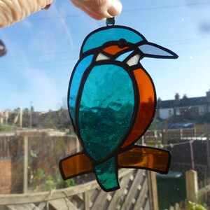 May include: A stained glass ornament of a blue, green, and orange kingfisher bird with a long beak. The bird is hanging from a green string.