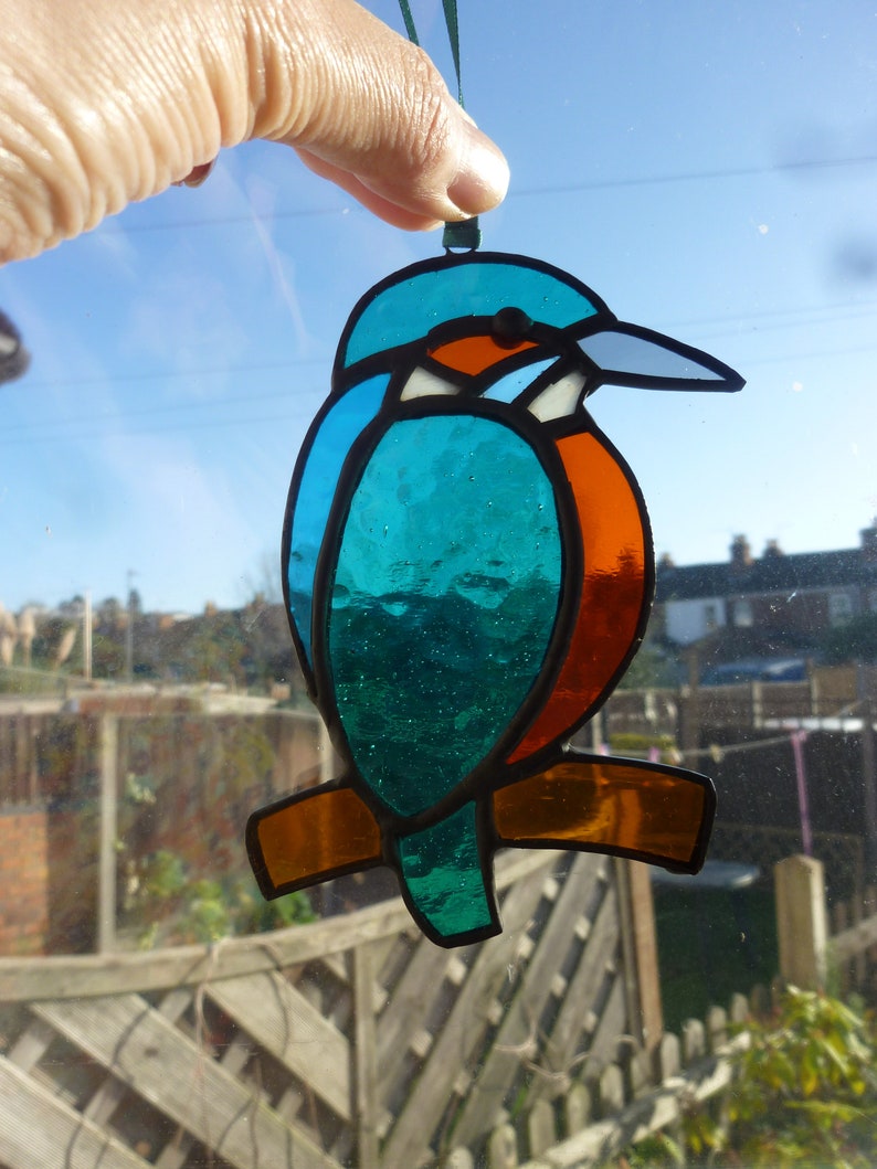 May include: A stained glass ornament of a blue, green, and orange kingfisher bird with a long beak. The bird is hanging from a green string.