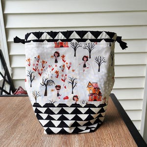 May include: A drawstring bag with a Halloween-themed print. The bag features a black and white triangle pattern at the top and bottom. The main body of the bag has a white background with a spooky illustration of houses, trees, and figures.