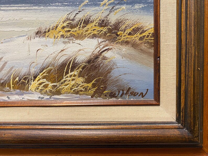 Op de afbeelding: Een close-up van een schilderij dat een besneeuwd landschap met hoog gras en een blauwe lucht afbeeldt. Het schilderij is rechtsonder getekend met "J. Wilson".