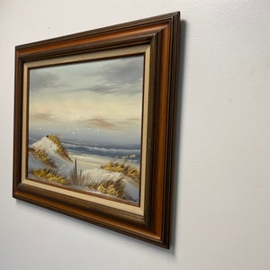 Op de afbeelding: Een ingelijst olieverfschilderij dat een zeezicht voorstelt met een zandstrand, duinen en een blauwe zee met witte toppen. De lucht is lichtblauw met witte wolken. Het schilderij is in een bruine houten lijst.