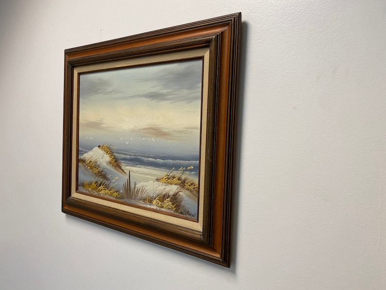 Op de afbeelding: Een ingelijst olieverfschilderij dat een zeezicht voorstelt met een zandstrand, duinen en een blauwe zee met witte toppen. De lucht is lichtblauw met witte wolken. Het schilderij is in een bruine houten lijst.