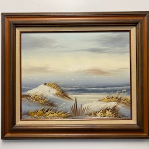 Op de afbeelding: Een ingelijst olieverfschilderij dat een zeezicht voorstelt met een zandstrand, golven die op de kust breken en een paar vogels die in de lucht vliegen. Het schilderij is geschilderd tegen een achtergrond van een bewolkte lucht met een vleugje gouden licht.