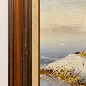 Op de afbeelding: Een ingelijst schilderij van een besneeuwd zeelandschap met hoog gras op de voorgrond. Het schilderij is in een donkerhouten lijst met een lichtbruine passe-partout.