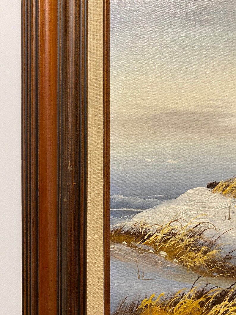 Op de afbeelding: Een ingelijst schilderij van een besneeuwd zeelandschap met hoog gras op de voorgrond. Het schilderij is in een donkerhouten lijst met een lichtbruine passe-partout.