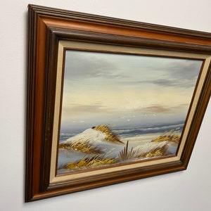 Op de afbeelding: Een ingelijst schilderij dat een zeezicht voorstelt met een zandstrand, zeegras en een paar vogels die overvliegen. Het schilderij is in een bruine houten lijst.