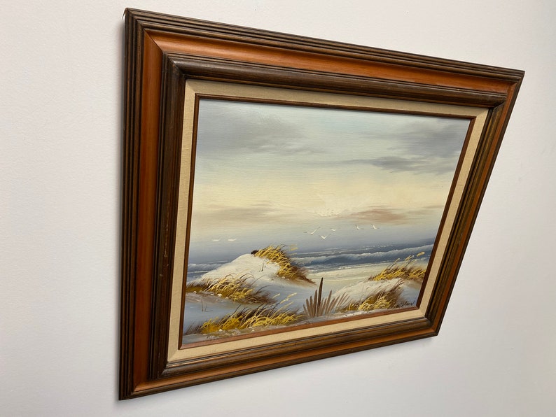 Op de afbeelding: Een ingelijst schilderij dat een zeezicht voorstelt met een zandstrand, zeegras en een paar vogels die overvliegen. Het schilderij is in een bruine houten lijst.
