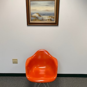 Op de afbeelding: Een feloranje stoel met een chromen onderstel staat op een grijs tapijt voor een witte muur met een ingelijst schilderij van een zeezicht.