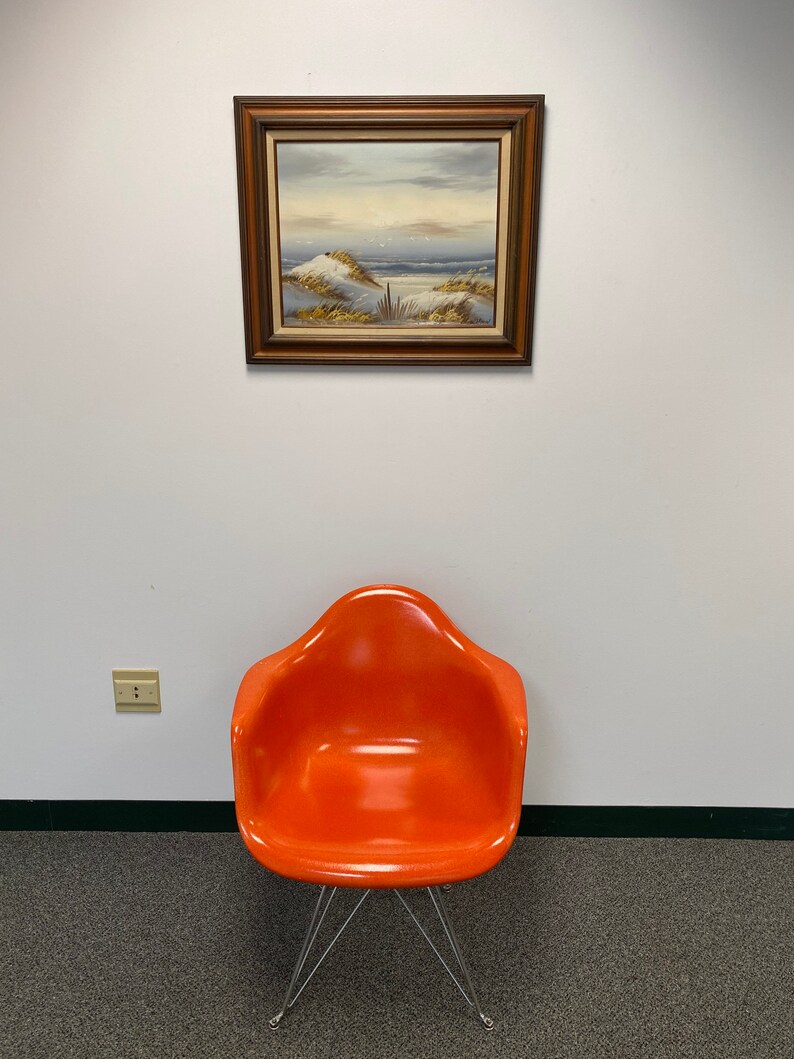 Op de afbeelding: Een feloranje stoel met een chromen onderstel staat op een grijs tapijt voor een witte muur met een ingelijst schilderij van een zeezicht.