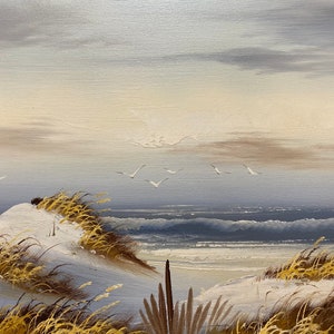 Op de afbeelding: Een schilderij van een zeelandschap met witte zandduinen, hoog gras en een blauwe zee met witte toppen. De lucht is lichtblauw met witte wolken.