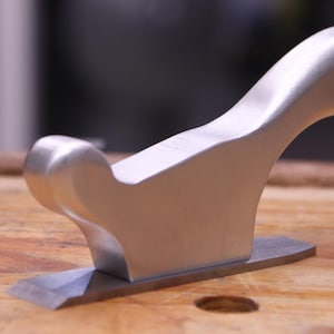May include: A silver-colored hand plane with a curved handle and a rectangular base. The blade is visible at the bottom. The tool is designed for woodworking and sits on a wooden surface.