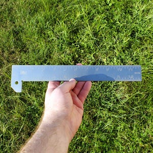 Puede incluir: Una regla de metal plateada con una escala negra marcada en pulgadas. La regla mide 12 pulgadas de largo y tiene un pequeño agujero en un extremo.