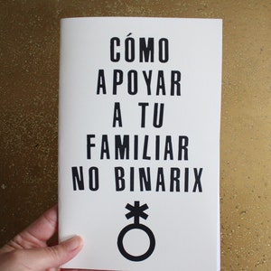 May include: A white booklet with the text "CÓMO APOYAR A TU FAMILIAR NO BINARIX" in black. A black and white gender-neutral symbol is below the text.