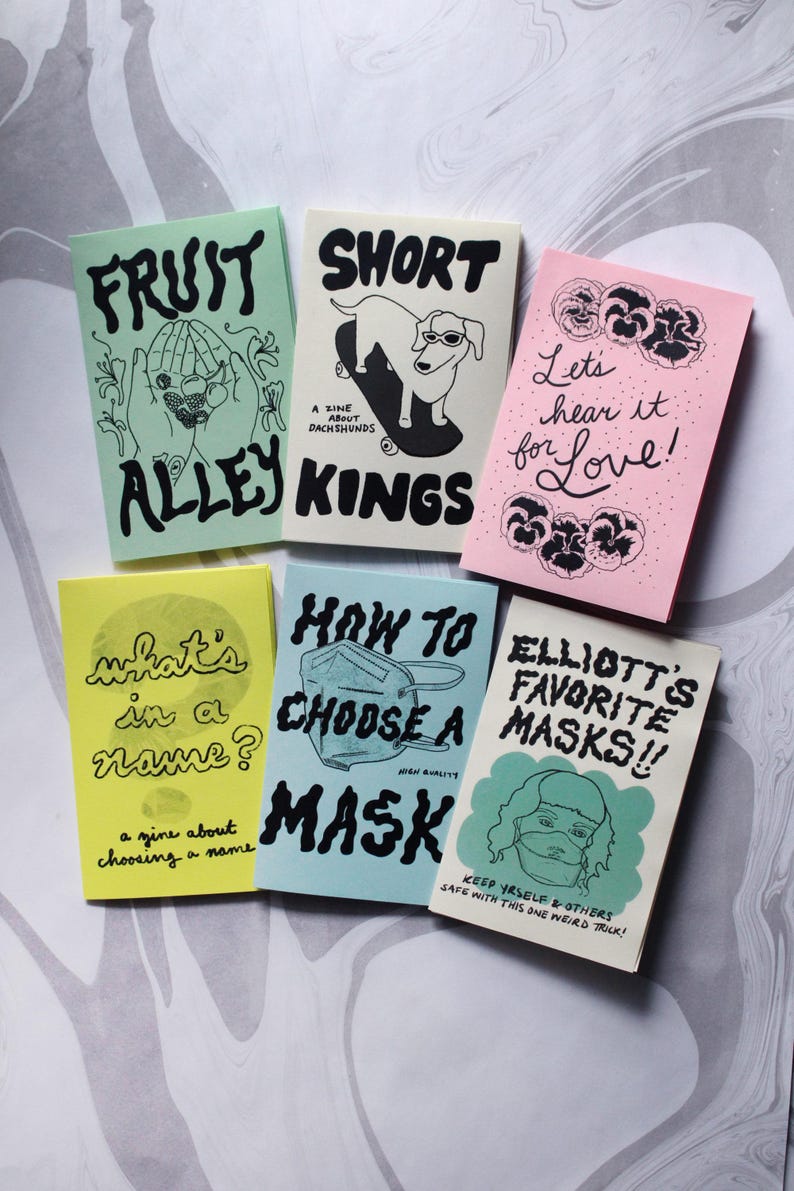May include: Six zines with colorful covers featuring black ink illustrations and text. Titles include Fruit Alley, Short Kings, Let's hear it for Love, what's in a name?, How to Choose a Mask, and Elliott's Favorite Masks, showcasing various graphic styles.