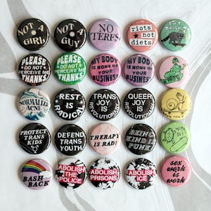 May include: A collection of twenty-five circular pin-back buttons arranged in a grid. The buttons feature various colors and designs, including text such as Not A Girl, No Terfs, Riots Not Diets, Normalize Acne, Trans Joy Is Revolutionary, and Abolish The Police.