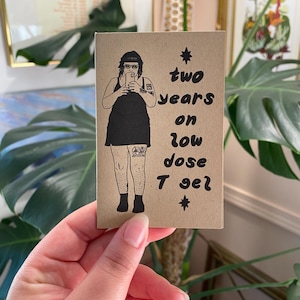 May include: A rectangular tan card featuring a black ink illustration of a person in a tank top and skirt taking a mirror selfie. The text reads: two years on low dose T gel, accompanied by two small star symbols, one above and one below the text.