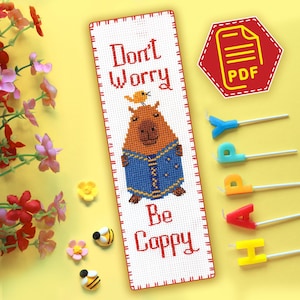 May include: White cross-stitch bookmark with a red border. The bookmark features a cartoon capybara wearing a blue denim jacket and a yellow bird perched on its head. The text "Don't Worry Be Cappy" is stitched in red.