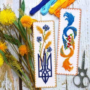 May include: Two embroidered bookmarks with a white background and orange borders. The bookmark on the left features a blue trident with wheat stalks and blue flowers. The bookmark on the right features a blue and orange bird with red berries.