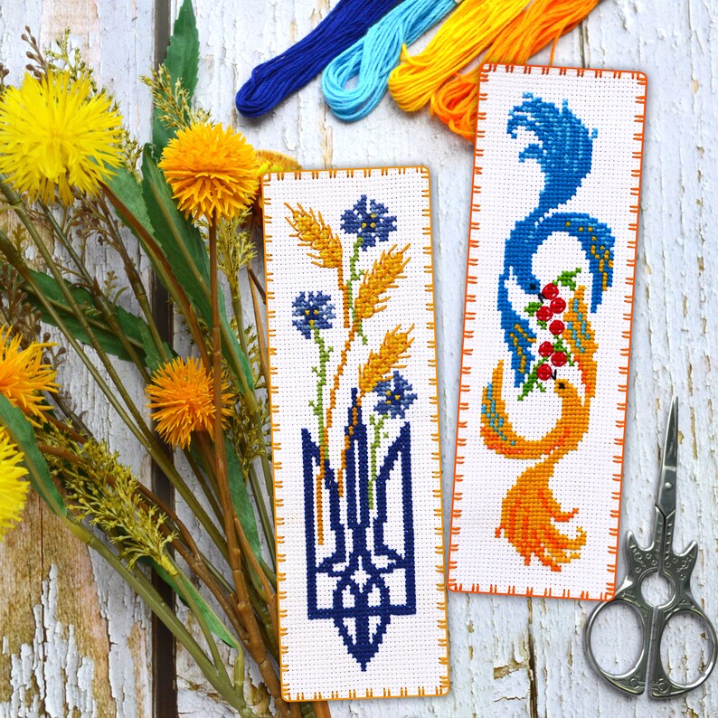 May include: Two embroidered bookmarks with a white background and orange borders. The bookmark on the left features a blue trident with wheat stalks and blue flowers. The bookmark on the right features a blue and orange bird with red berries.