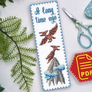 Puede incluir: Un marcapáginas de punto de cruz con un borde azul y el texto "A long time ago" en letras azules. El marcapáginas presenta una escena de montaña con dos pterodáctilos volando por encima. El marcapáginas es una descarga PDF.