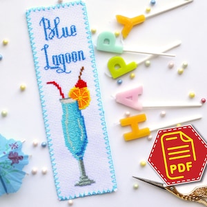 May include: A cross-stitch bookmark with a blue lagoon cocktail design. The bookmark is white with blue, orange, and red stitching. The text "Blue Lagoon" is stitched in black thread. The bookmark is surrounded by colorful sprinkles and a small blue umbrella.