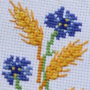 May include: Cross-stitch embroidery design featuring two yellow wheat stalks and two blue flowers with yellow centers. The design is on a white background.