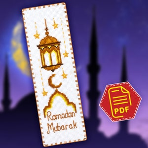 Può includere: Un segnalibro bianco a punto croce con una lanterna dorata, una mezzaluna e stelle. Il testo "Ramadan Mubarak" è ricamato in filo marrone.
