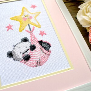 K&ouml;nnte beinhalten: Eine Kreuzstich-Stickerei mit einem Pandab&auml;ren, der einen gelben Stern mit einem l&auml;chelnden Gesicht h&auml;lt. Der Panda tr&auml;gt ein rosa-wei&szlig; gestreiftes Outfit und ist von kleinen rosa Sternen umgeben.