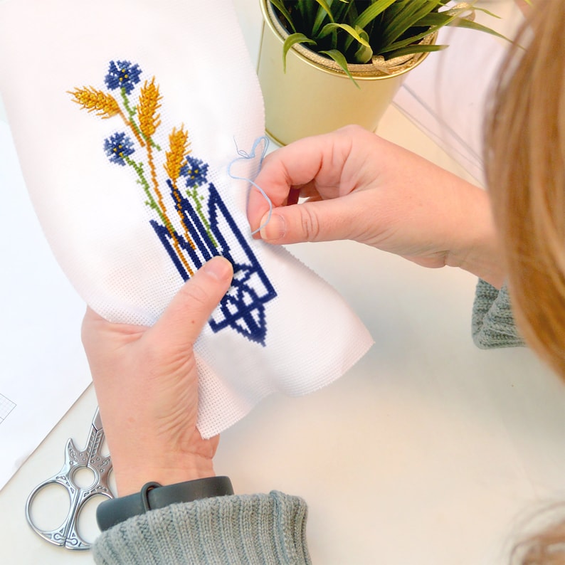 May include: A person cross-stitching a white fabric with a blue and yellow design. The design features a stylized trident with wheat stalks and blue flowers.