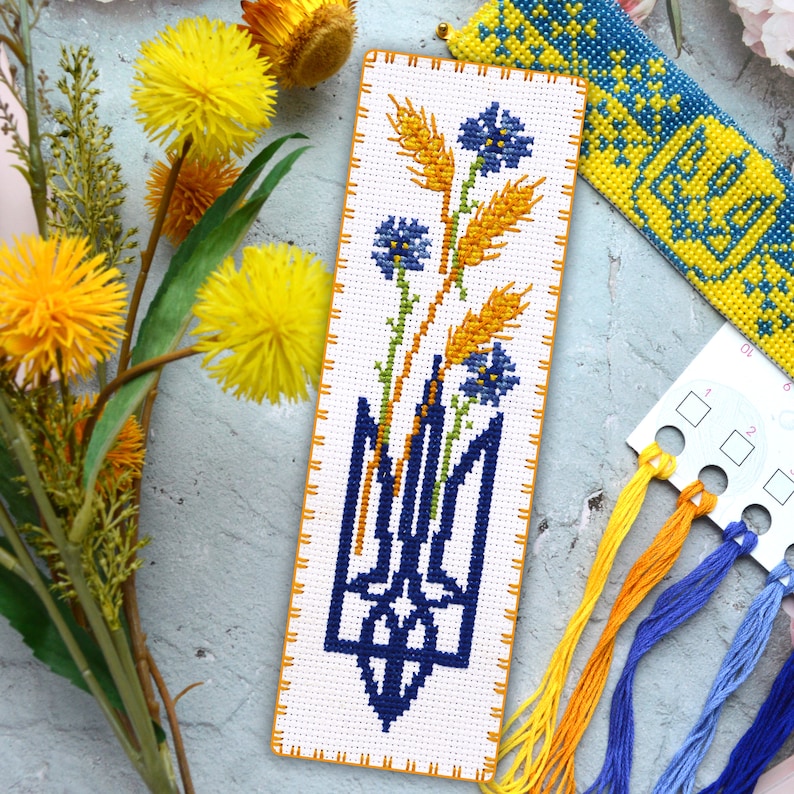 May include: A cross-stitch bookmark featuring a blue Ukrainian trident with wheat stalks and blue flowers. The bookmark is white with a yellow border.