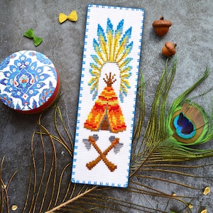 May include: A cross-stitch pattern of a Native American teepee with a colorful design. The teepee is surrounded by feathers and has a yellow and orange design. The pattern is on white fabric with a blue border.